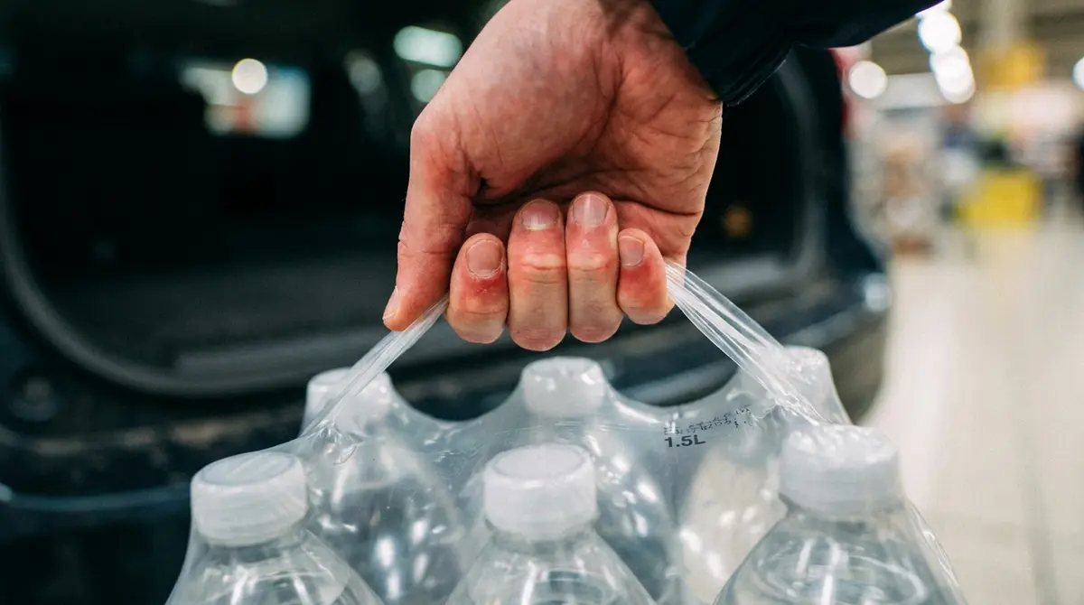 Primer plano de manos sufriendo al cargar un pack pesado de botellas de agua tradicionales en plástico termoencogible.